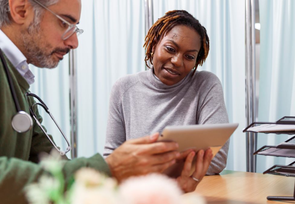An image of a woman with brown skin and short brown braids, wearing a grey sweater, talking to a male doctor with grey hair, a gray beard, white skin, glasses, a green sweater, and wearing a stethoscope. They are reviewing a tablet together and the image leads to a Milken Institute article around a preventative health roundtable. 